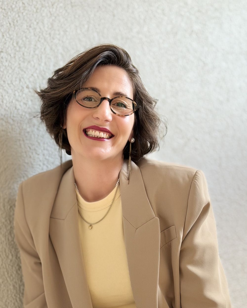 Patricia lacht, im Blazer an eine Wand gelehnt